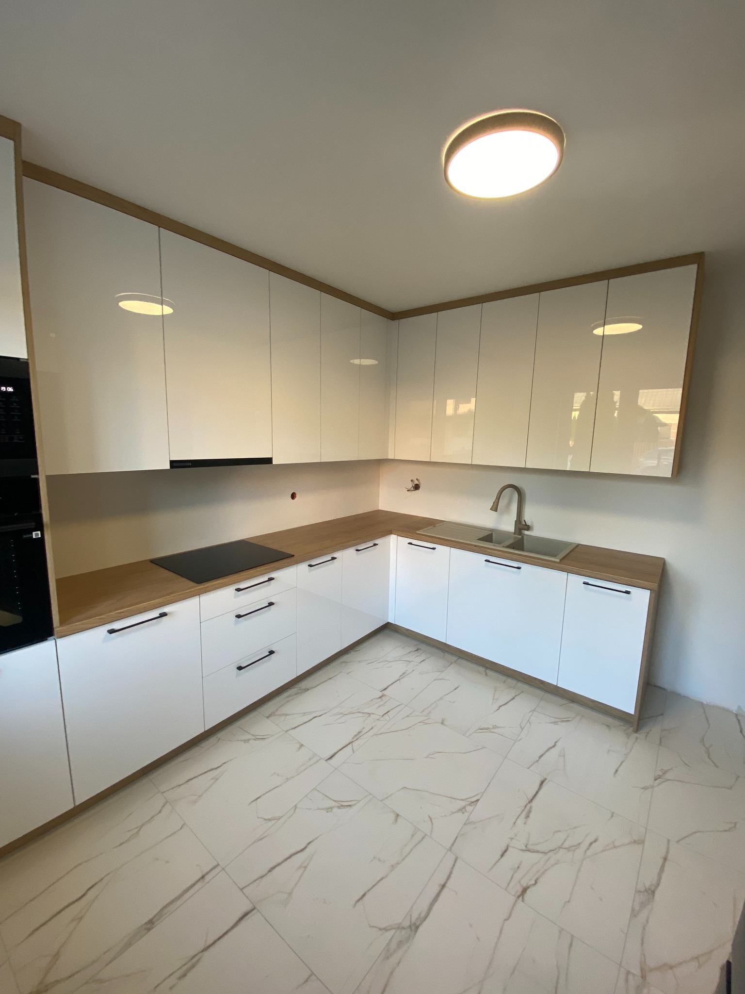 Nowoczesna, narożna kuchnia z białymi, lakierowanymi frontami i drewnianymi blatami, czarnymi, podłużnymi uchwytami i marmurową podłogą