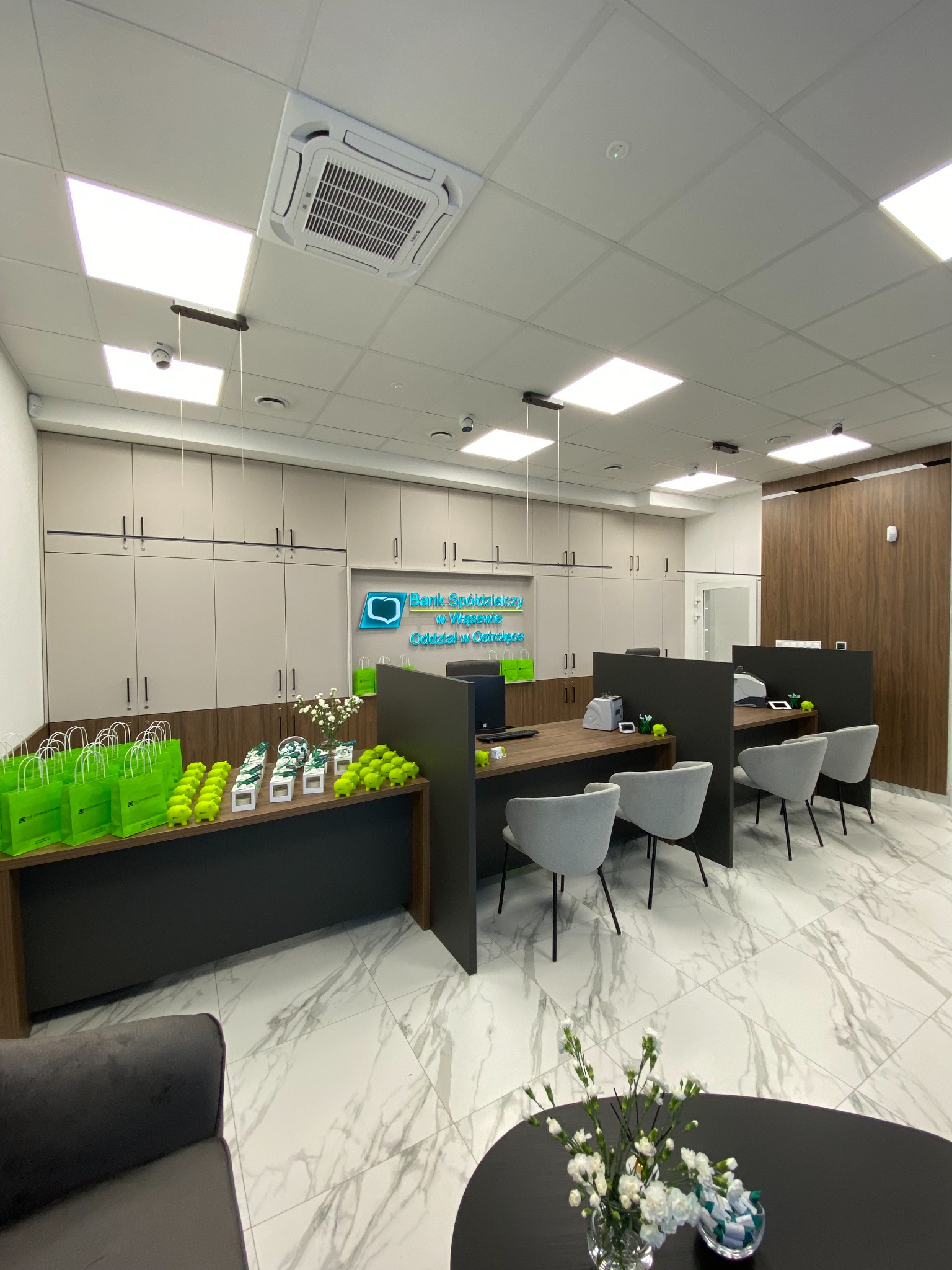 Wnętrze nowocześnie urządzonego oddziału banku z biurkami, szarymi krzesłami, szafkami w tle, jasną podłogą z marmurowym wzorem oraz zielonymi torbami i figurkami na blacie.
