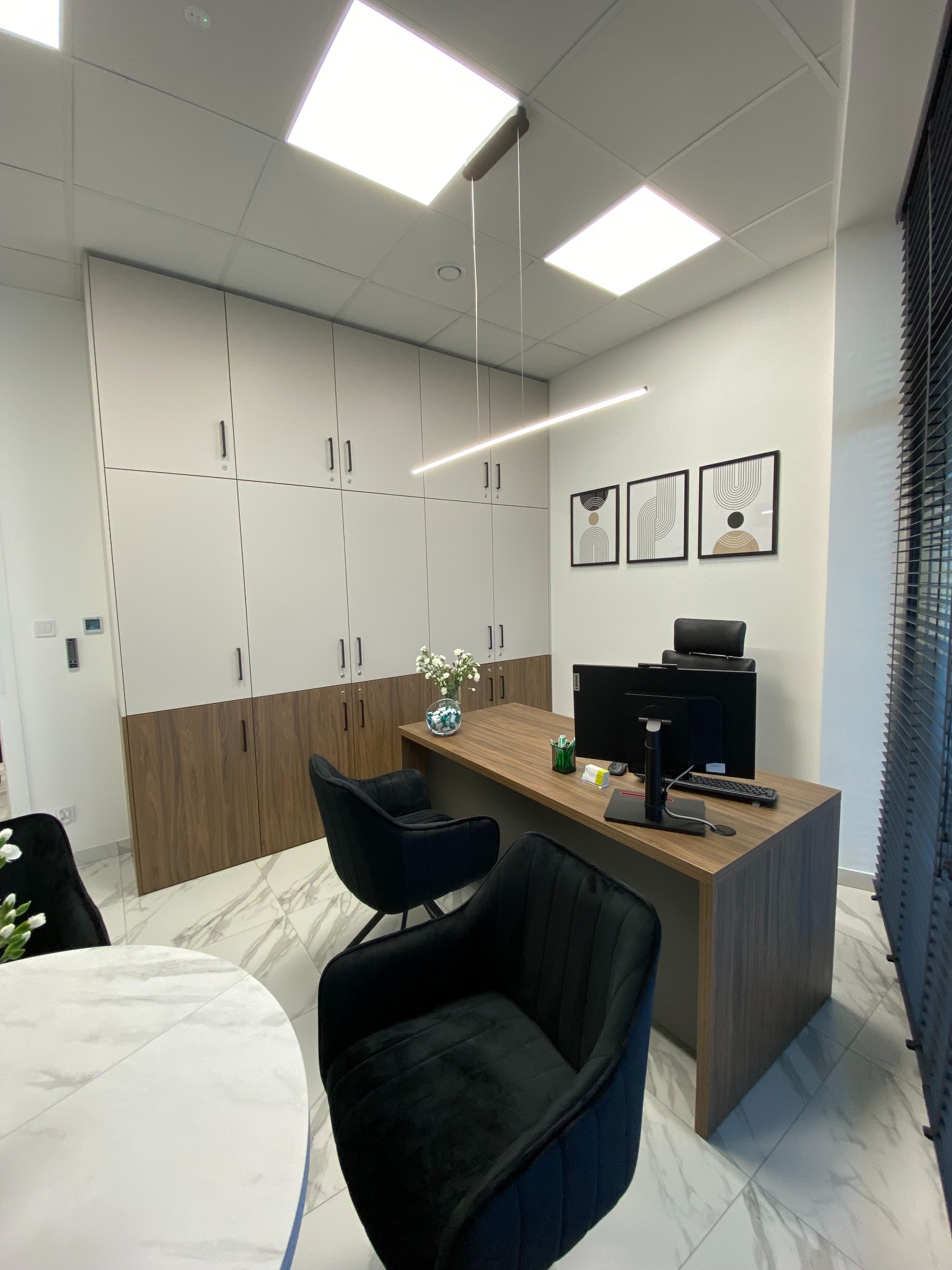 Nowoczesne biuro z drewnianym biurkiem, czarnymi krzesłami welurowymi, białymi szafkami z drewnianymi akcentami, minimalistycznymi obrazami na ścianie i marmurową podłogą.