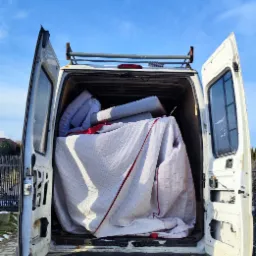 Otwarty tył białego busa z załadowanymi poduszkami i materacem przykrytymi białą kołdrą w czerwoną kratę, na brukowanym podjeździe pod błękitnym niebem.