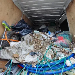 Wnętrze furgonetki wypełnionej gruzem, odpadami budowlanymi i starymi rurami, zabezpieczonymi zieloną taśmą transportową, widok z tyłu pojazdu w Poznaniu.