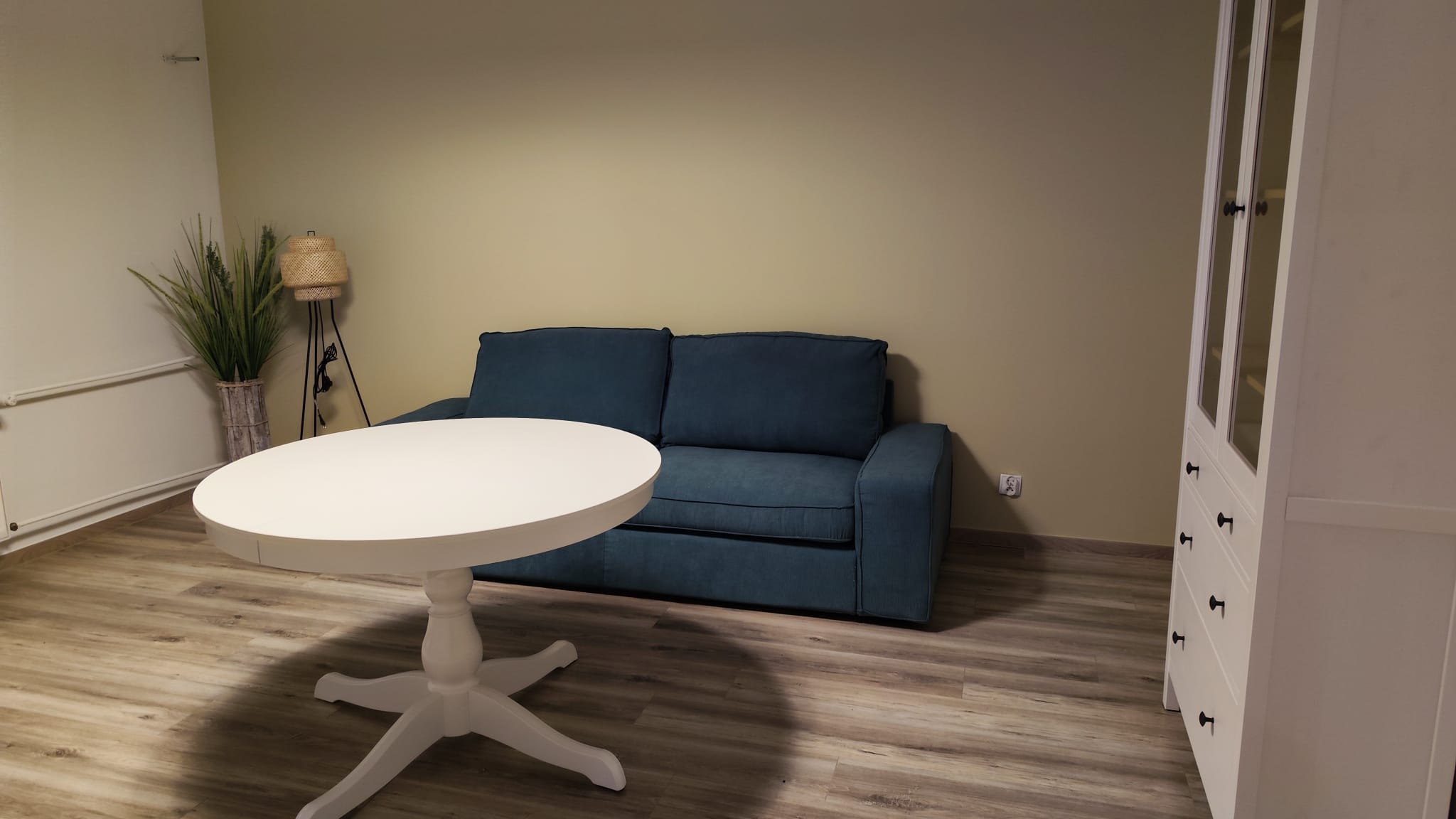 Narożnik w odcieniach niebieskiego, biały okrągły stół na nodze z krzyżowym podparciem, biała komoda z przeszkloną nadstawką, lampa z abażurem z rattanu i wysoka roślina w ozdobnej donicy...