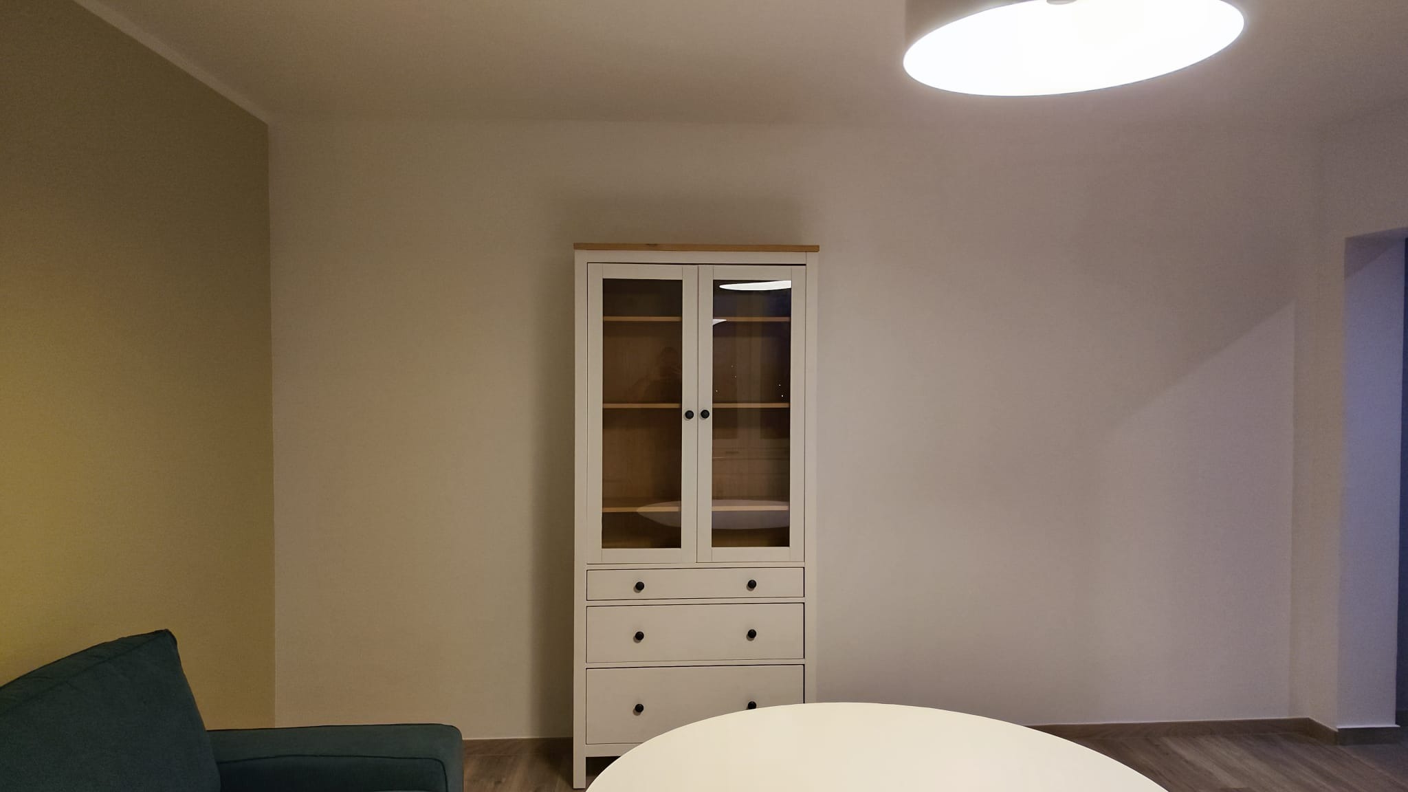 Biała witryna IKEA z drewnianym blatem, trzema szufladami i przeszkloną górną częścią, stojąca na drewnianej podłodze w minimalistycznym salonie z kremowymi ścianami i fragmentem zielonej sofy.