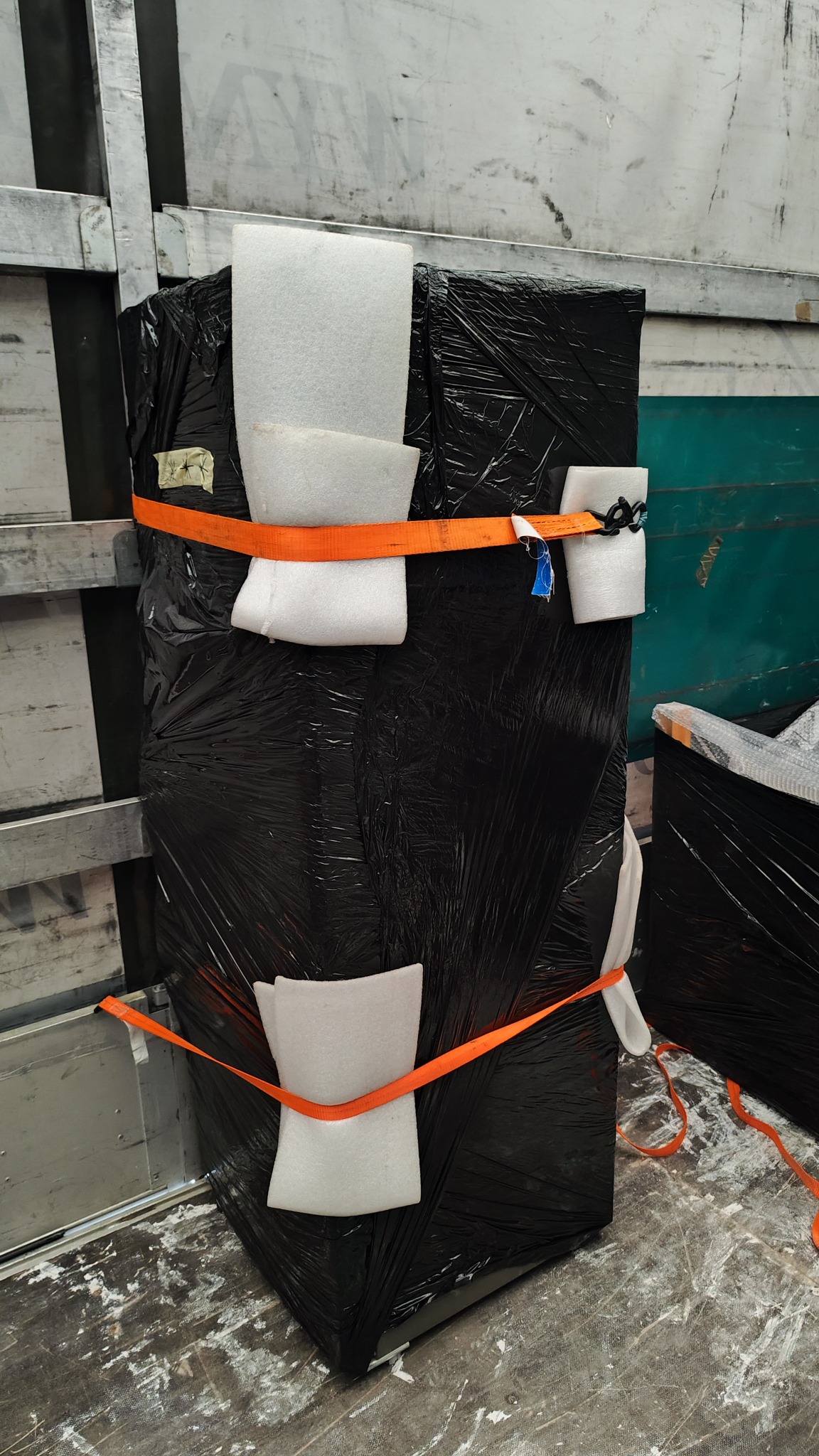 Zabezpieczony pakunek owinięty czarną folią i pianką, przymocowany pomarańczowymi pasami transportowymi wewnątrz pojazdu dostawczego.