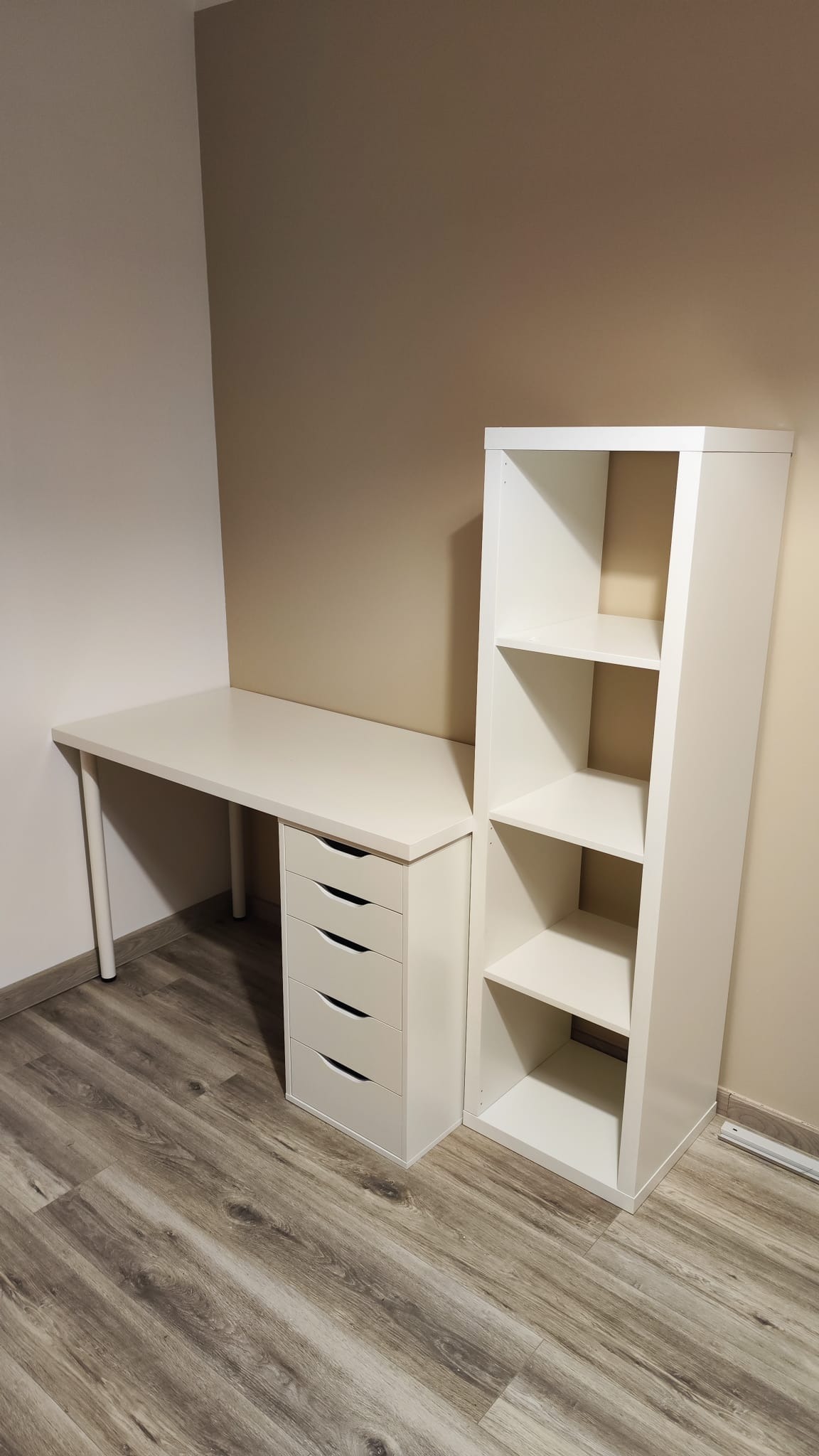 Białe biurko z szufladami i regałem z półkami, ustawione na podłodze imitującej drewno, na tle dwukolorowej ściany.