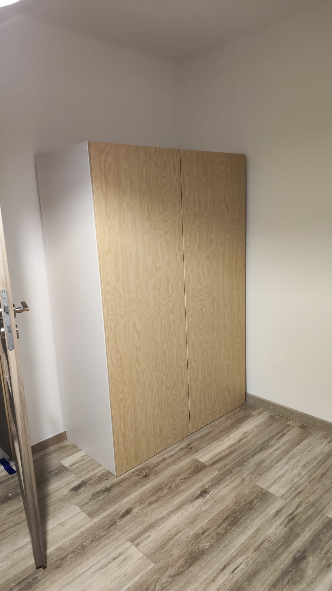 Zmontowana szafa IKEA z jasnego drewna z widocznym usłojeniem, stojąca w rogu pomieszczenia z panelami podłogowymi w odcieniach szarości i beżu, widoczna klamka drzwi po lewej stronie.