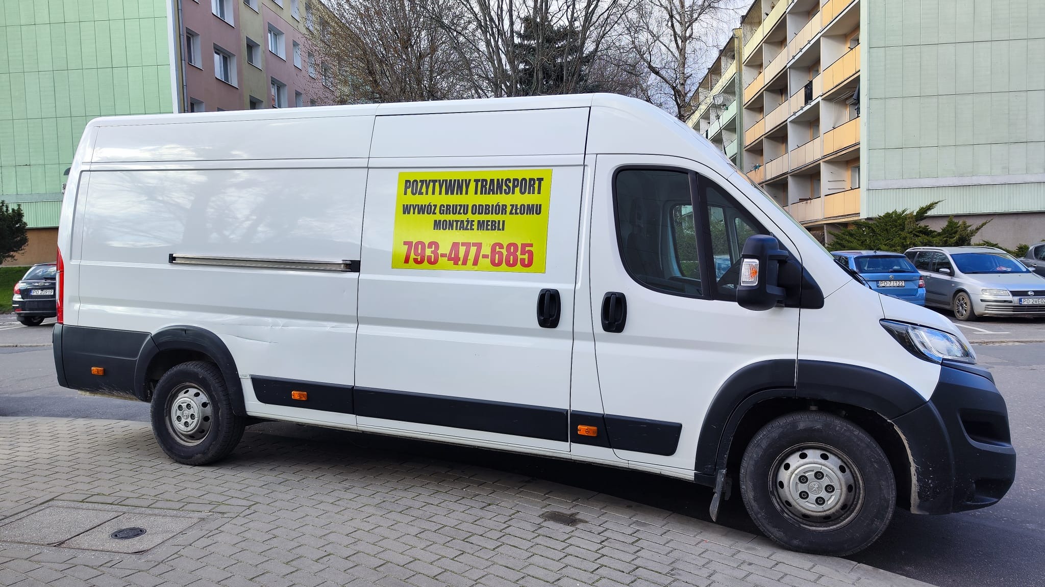 Biały samochód dostawczy z żółtą reklamą firmy 'Pozytywny Transport', oferującej wywóz gruzu, odbiór złomu i montaż mebli, zaparkowany na chodniku na tle bloków mieszkalnych.