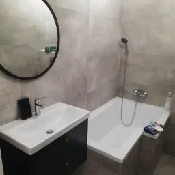 Nowoczesna łazienka w szarościach z okrągłym lustrem, umywalką z czarną szafką i białą wanną z baterią.