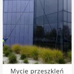 Pracownik w niebieskiej kurtce myje szklaną fasadę budynku za pomocą długiej tyczki z szczotką; widoczne smugi wody.
