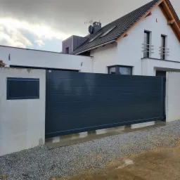 Nowoczesna, przesuwna brama w kolorze antracytowym, z poziomymi panelami, zamontowana przed białym domem z szarą dachówką, widoczne kamyczki na podłożu.