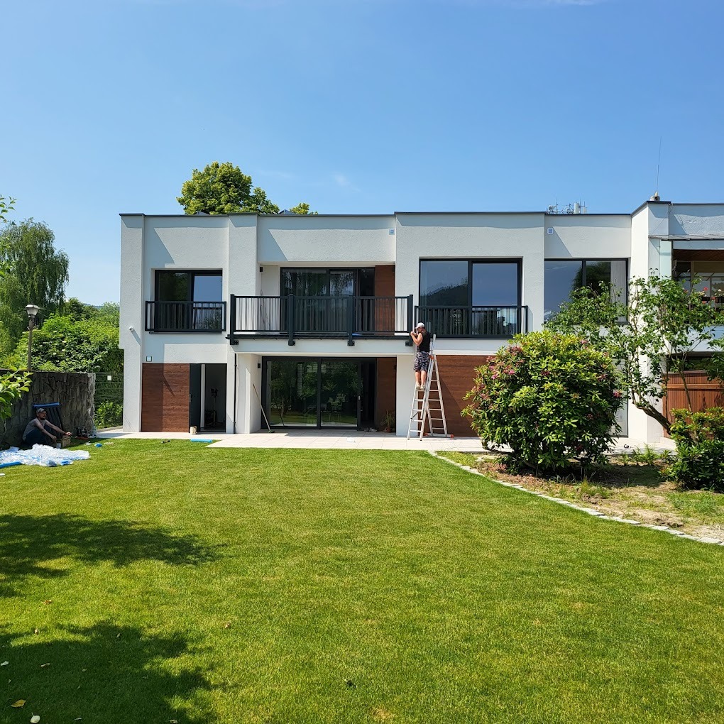 Pracownik na drabinie czyści okno na piętrze nowoczesnego domu z dużym trawnikiem, w tle widoczna osoba siedząca na pniu drzewa.