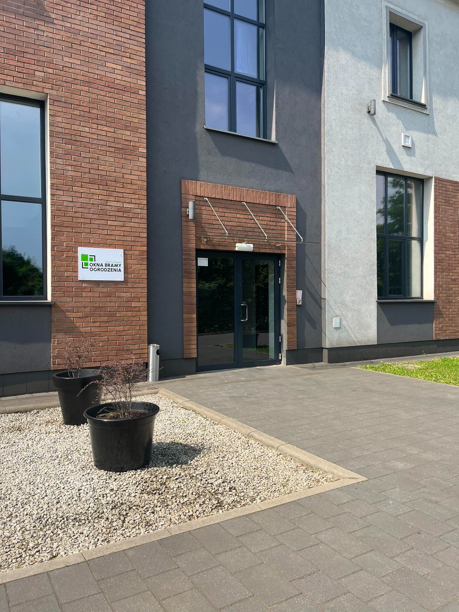 Fasada budynku z oknami PCV i drzwiami wejściowymi, z widocznym szyldem firmy 'Okna Bramy Ogrodzenia' oraz dwoma donicami z roślinami przed wejściem.