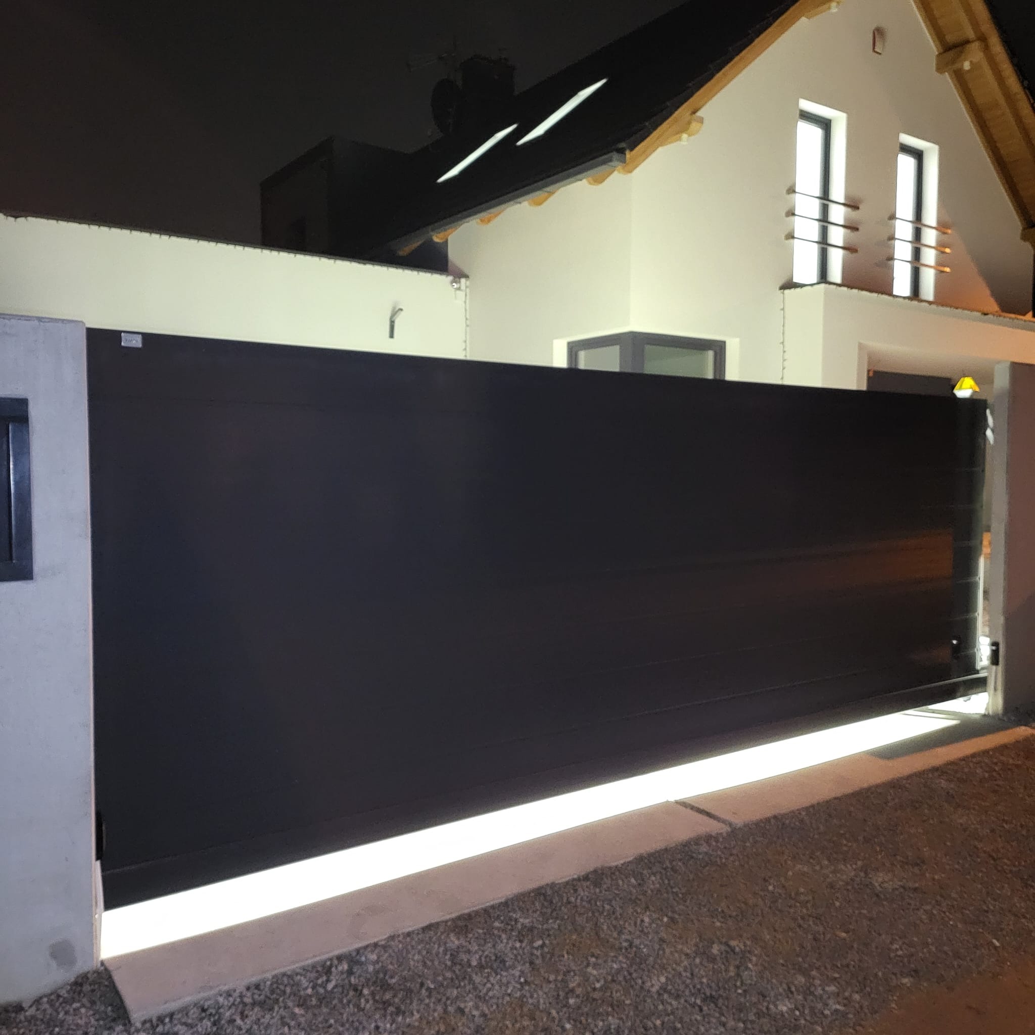 Czarna, przesuwna brama wjazdowa z podświetleniem LED u dołu, na tle oświetlonego domu jednorodzinnego nocą.