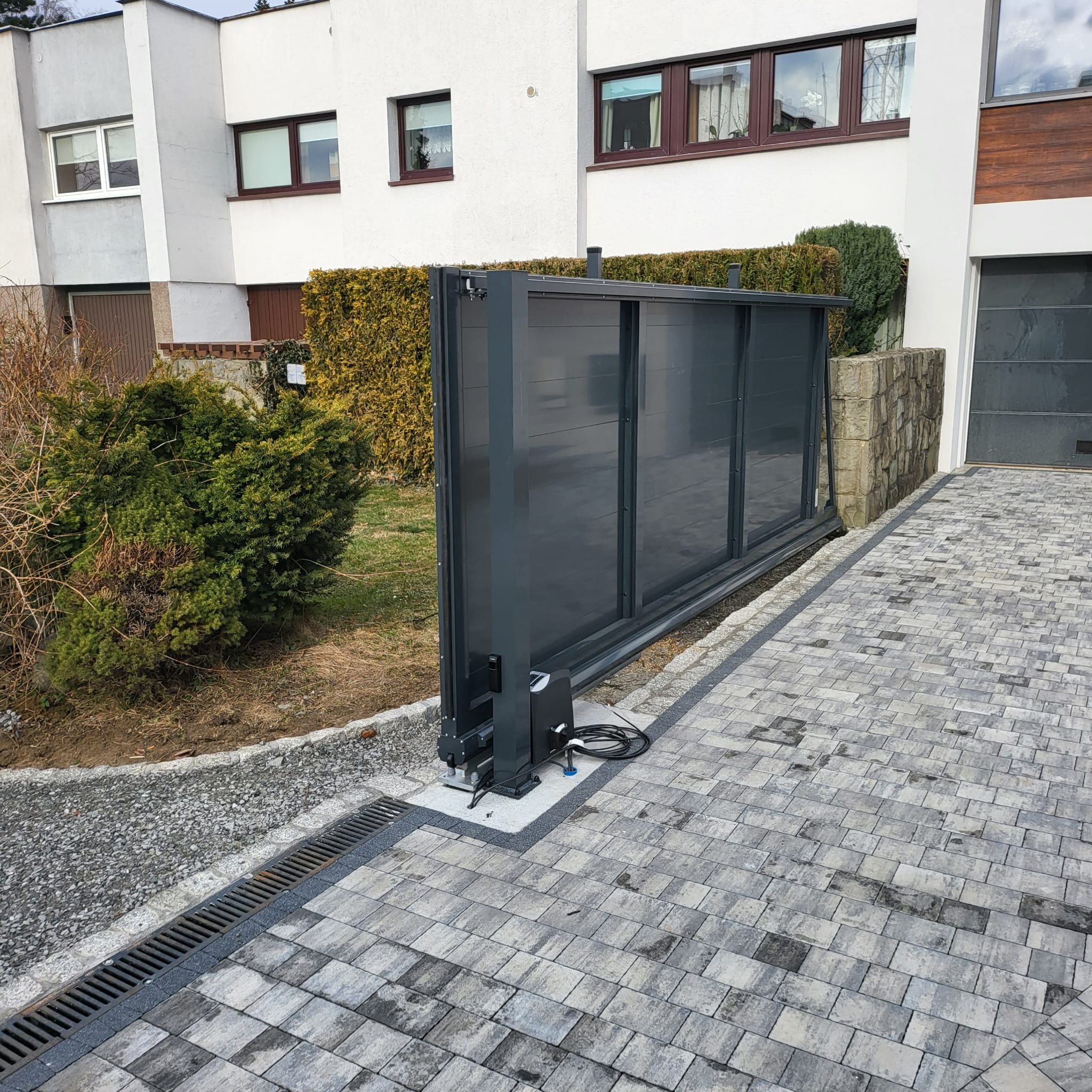 Automatyczna brama przesuwna w kolorze grafitowym, zamontowana na posesji z kostki brukowej, widoczny mechanizm napędowy i przewody elektryczne, w tle nowoczesny dom z białymi ścianami i brązowymi...