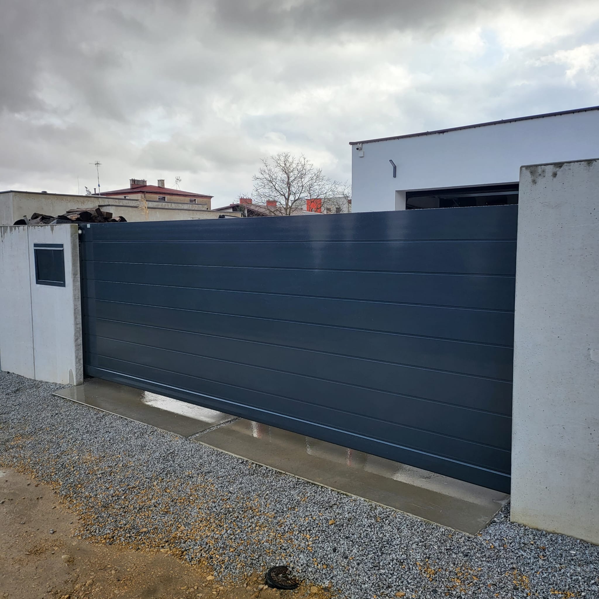 Nowoczesna, przesuwna brama wjazdowa w kolorze grafitowym z poziomymi panelami, zamontowana między betonowymi słupkami na posesji z szarym żwirem, pochmurne niebo w tle.