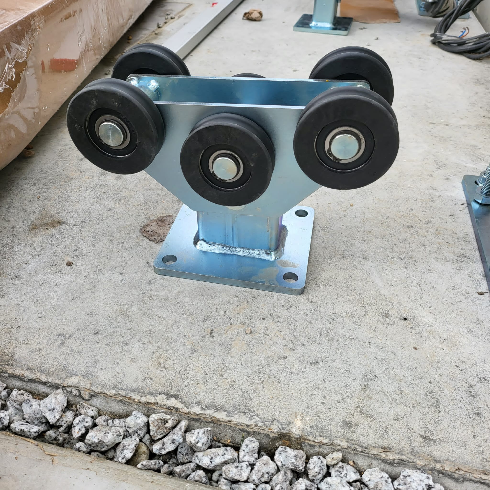 Wózek jezdny bramy przesuwnej z sześcioma czarnymi rolkami na metalowej konstrukcji, widoczne spawy mocujące, na betonowej posadzce z obrzeżem z białego grysu.