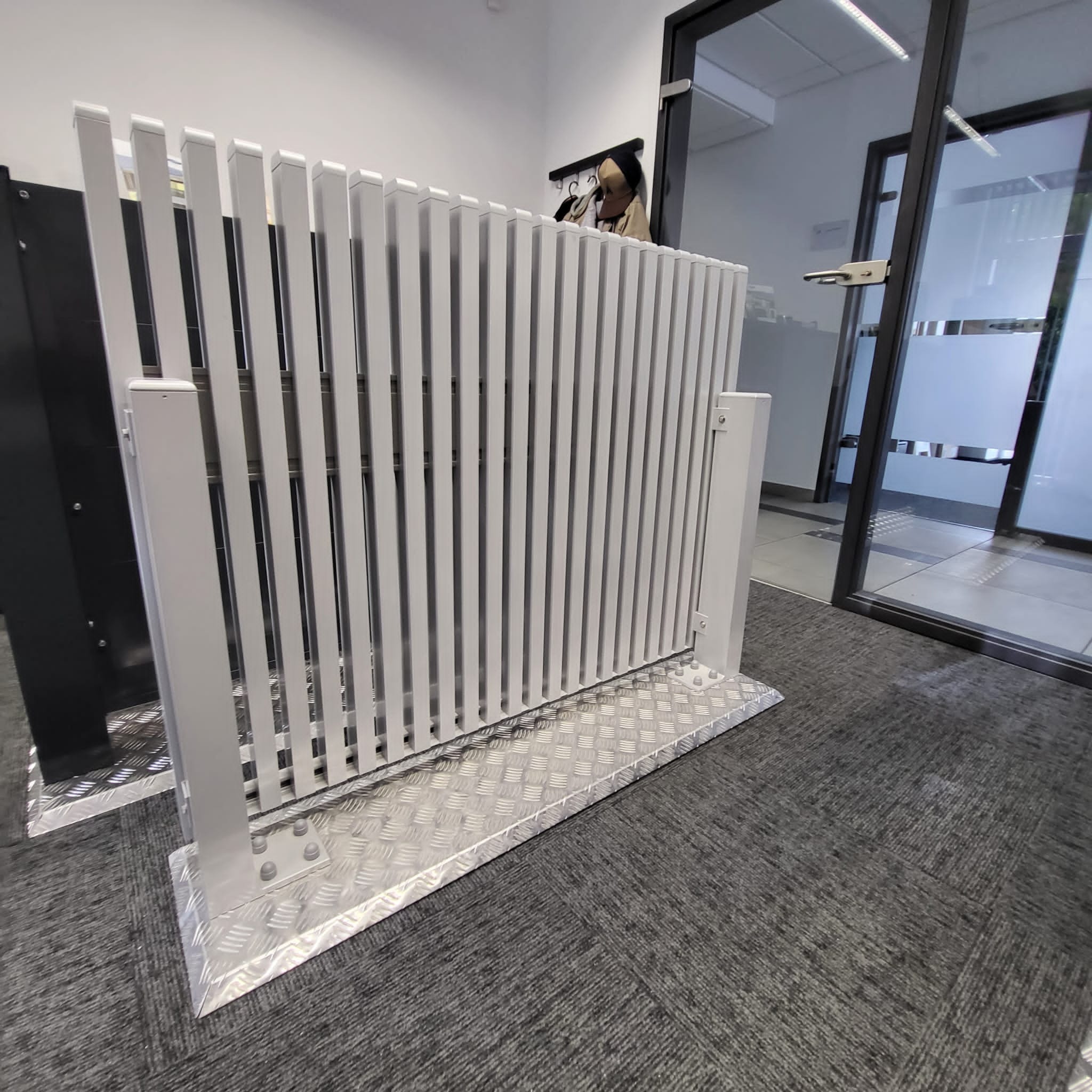 Biała, metalowa barierka ochronna z ryflowaną podstawą, zamontowana wewnątrz budynku, widoczna na tle szklanych drzwi i szarej wykładziny podłogowej.