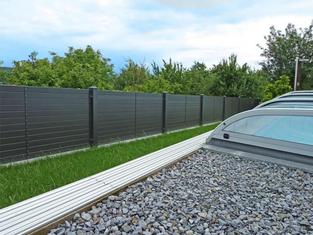 Nowoczesne, poziome ogrodzenie panelowe w kolorze grafitowym, oddzielające trawnik od terenu z szarym żwirem i częściowo widoczną konstrukcją zadaszenia basenu.