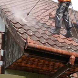 Pracownik w pomarańczowej kurtce i czarnych spodniach czyści dach z brudu przy użyciu myjki ciśnieniowej. Widoczna brudna dachówka ceramiczna i spływająca woda.