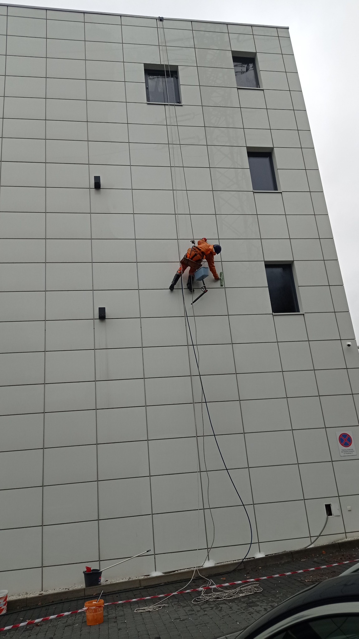 Pracownik w pomarańczowym kombinezonie, zabezpieczony linami, czyści elewację budynku z płytek, używając myjki i wiadra zawieszonego na uprzęży.