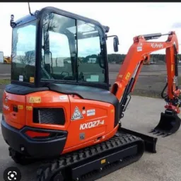 Pomarańczowa mini koparka Kubota KX027-4 na gąsienicach, zaparkowana na betonowej powierzchni, widok z boku, na tle zieleni i błękitnego nieba odbijającego się w szybach kabiny.