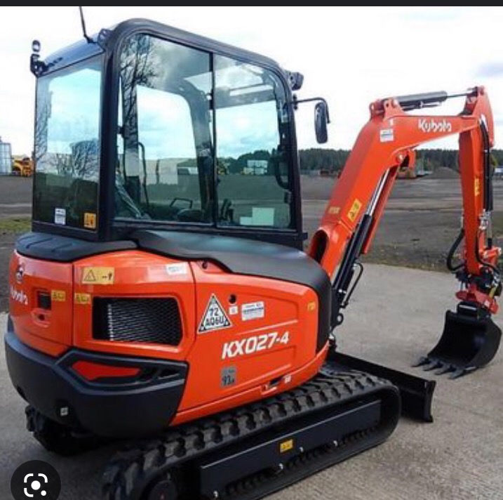 Pomarańczowa mini koparka Kubota KX027-4 na gąsienicach, zaparkowana na betonowej powierzchni, widok z boku, na tle zieleni i błękitnego nieba odbijającego się w szybach kabiny.