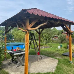 Drewniana altana z czerwoną dachówką w ogrodzie, widoczna drabina, stół z narzędziami i basen w tle.