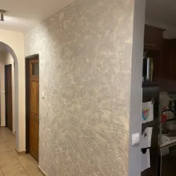 Ściana z dekoracyjną gładzią w odcieniach szarości i beżu, widoczny fragment korytarza z brązowymi drzwiami i fragment kuchni z lodówką ozdobioną rysunkami.