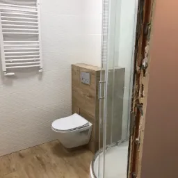 Nowoczesna łazienka w trakcie wykańczania: podwieszana toaleta, grzejnik drabinkowy na ścianie z trójwymiarowymi płytkami, kabina prysznicowa z widocznym surowym obramowaniem.