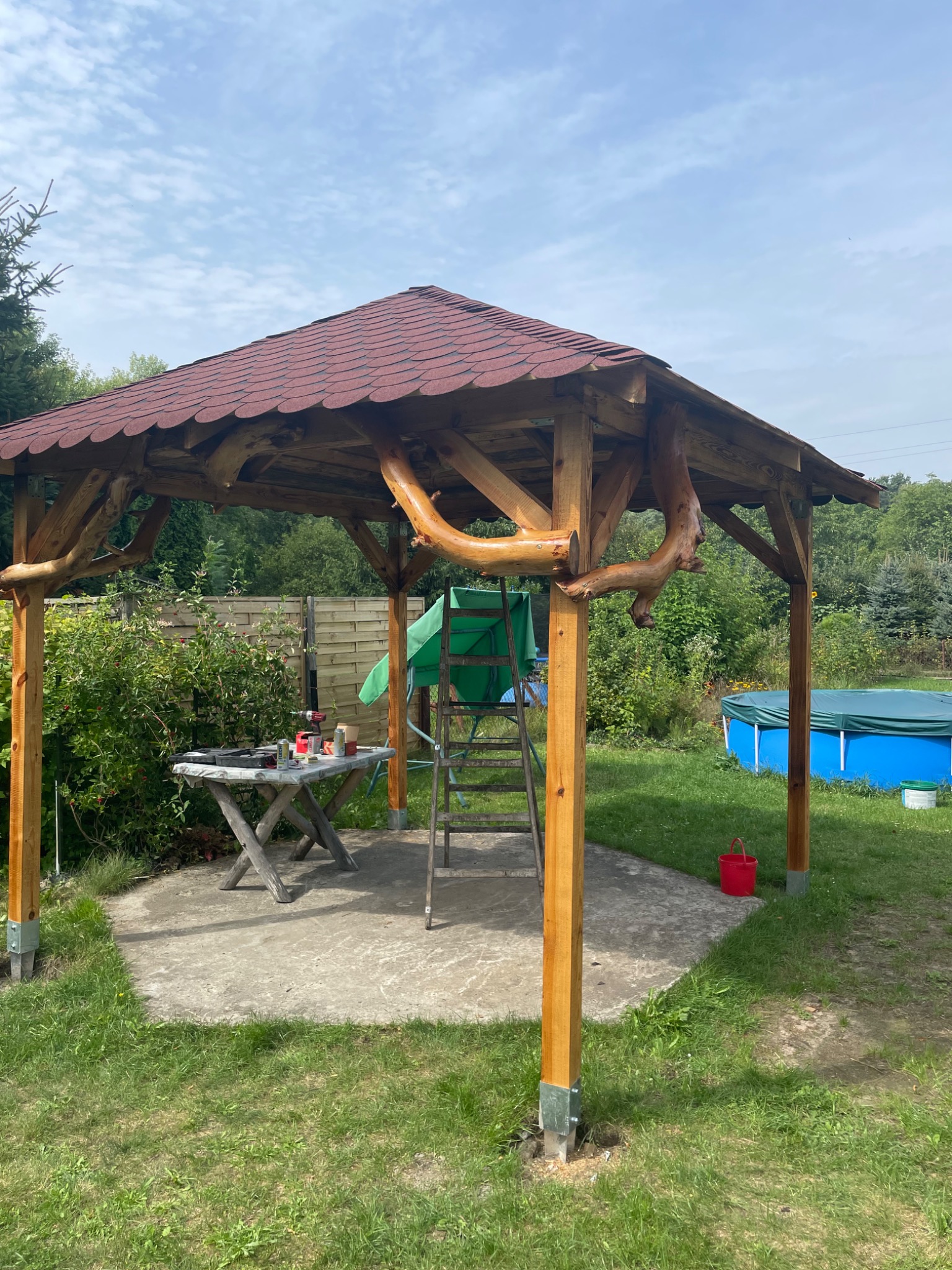 Drewniana altana z czerwoną dachówką w ogrodzie, z widocznym stołem roboczym, drabiną i basenem w tle, w trakcie budowy lub wykończenia.