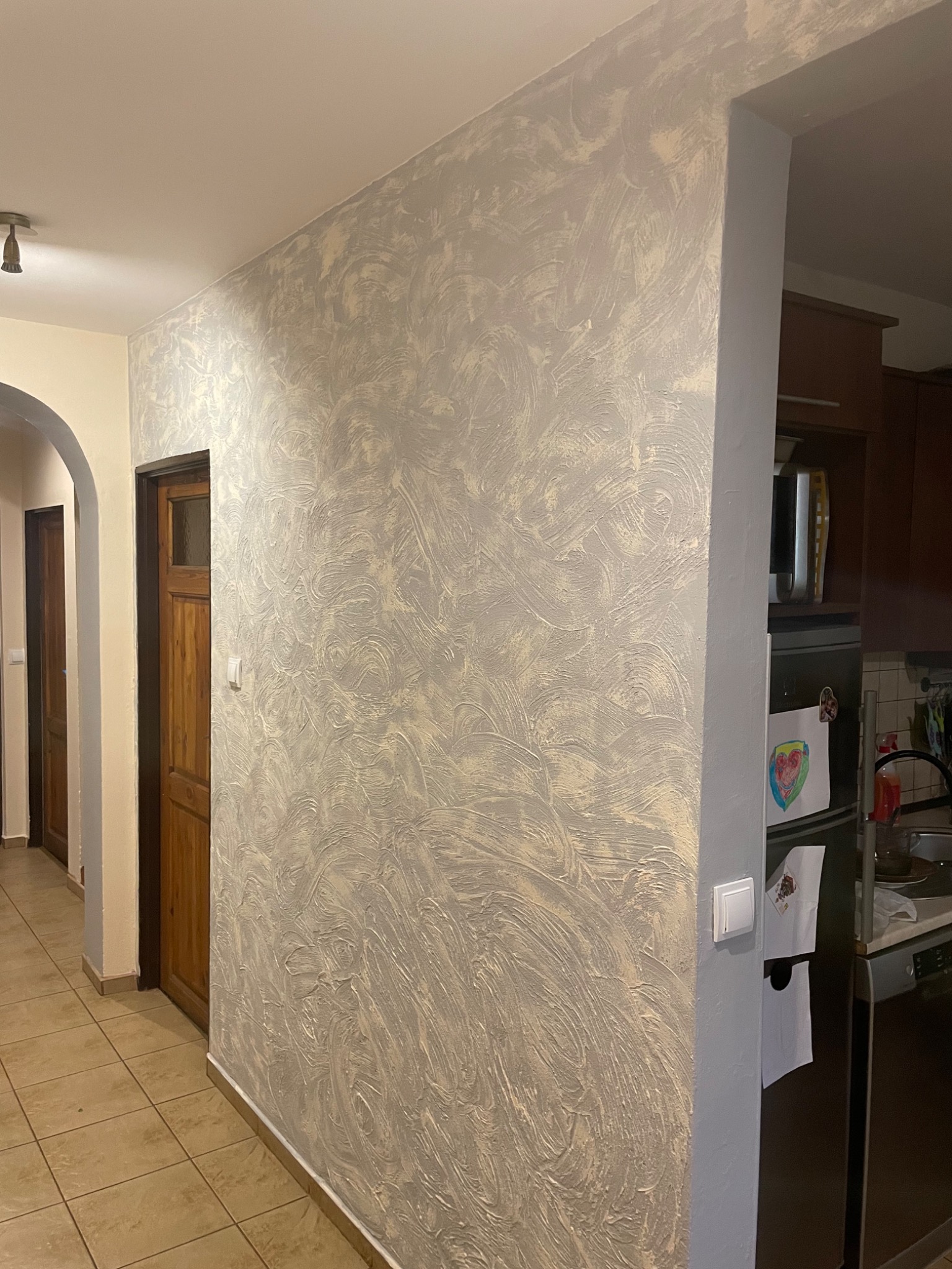 Ściana z dekoracyjną gładzią w odcieniach szarości i beżu, widoczny fragment korytarza z brązowymi drzwiami i fragment kuchni z lodówką ozdobioną rysunkami.