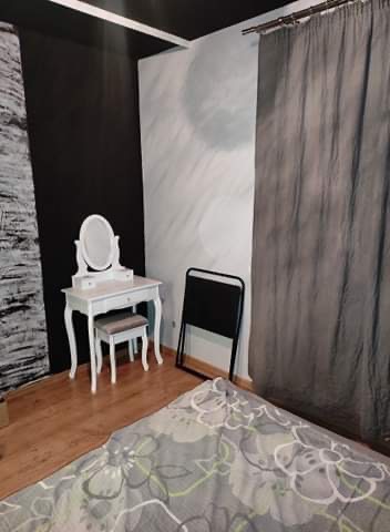 Ustawiony w rogu pokoju biały, toaletkowy stół z owalnym lustrem i tapicerowanym stołkiem, obok czarne składane krzesło i szara zasłona.