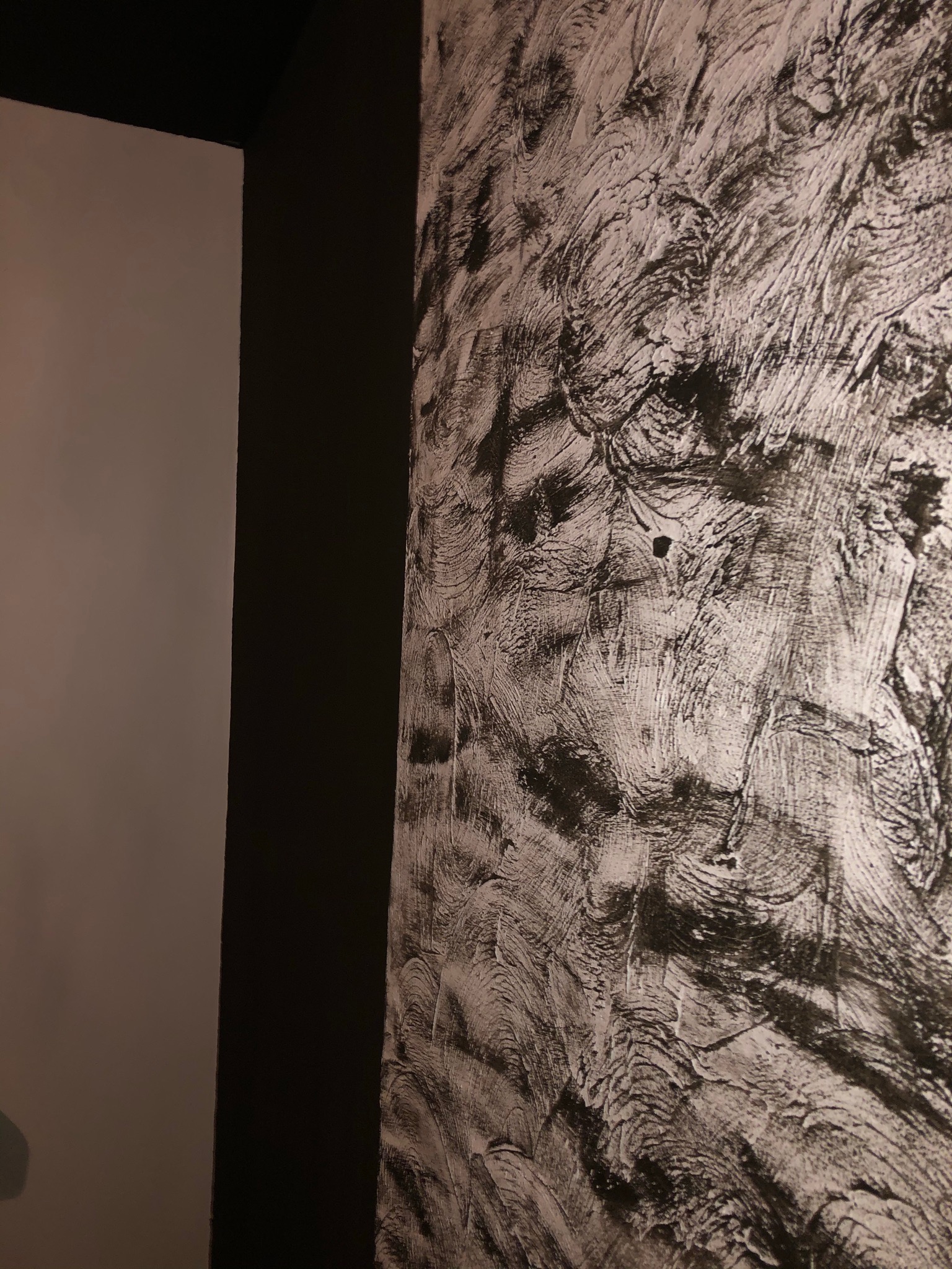 Fragment ściany z dekoracyjną, strukturalną gładzią w odcieniach szarości i bieli, sąsiadującej z gładką, ciemną ścianą i jasną ścianą w tle.