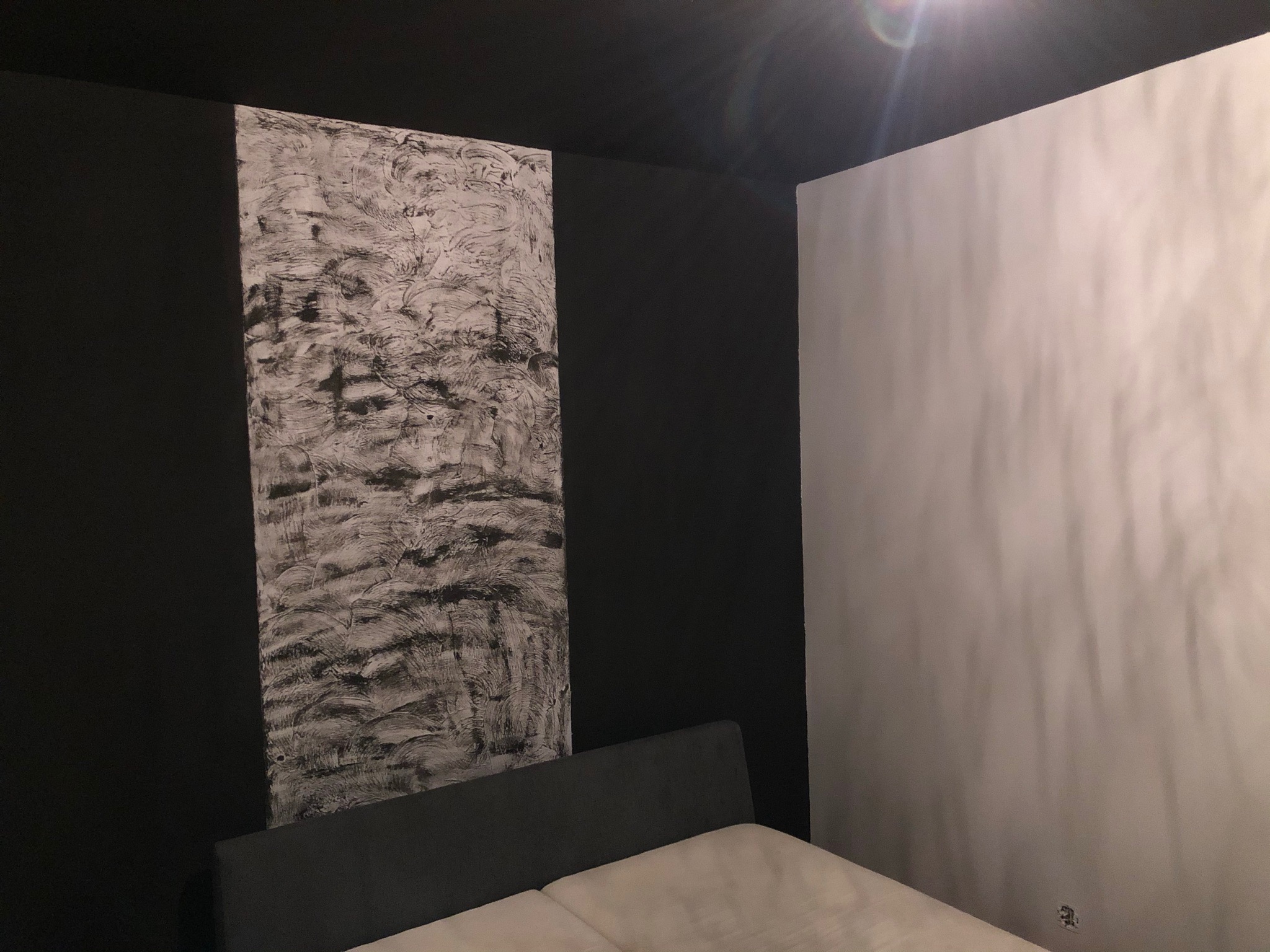 Sypialnia z dekoracyjną ścianą w odcieniach szarości, imitującą fakturę gładzi, kontrastująca z czarną ścianą i sufitem oraz białą ścianą, widoczny fragment łóżka.