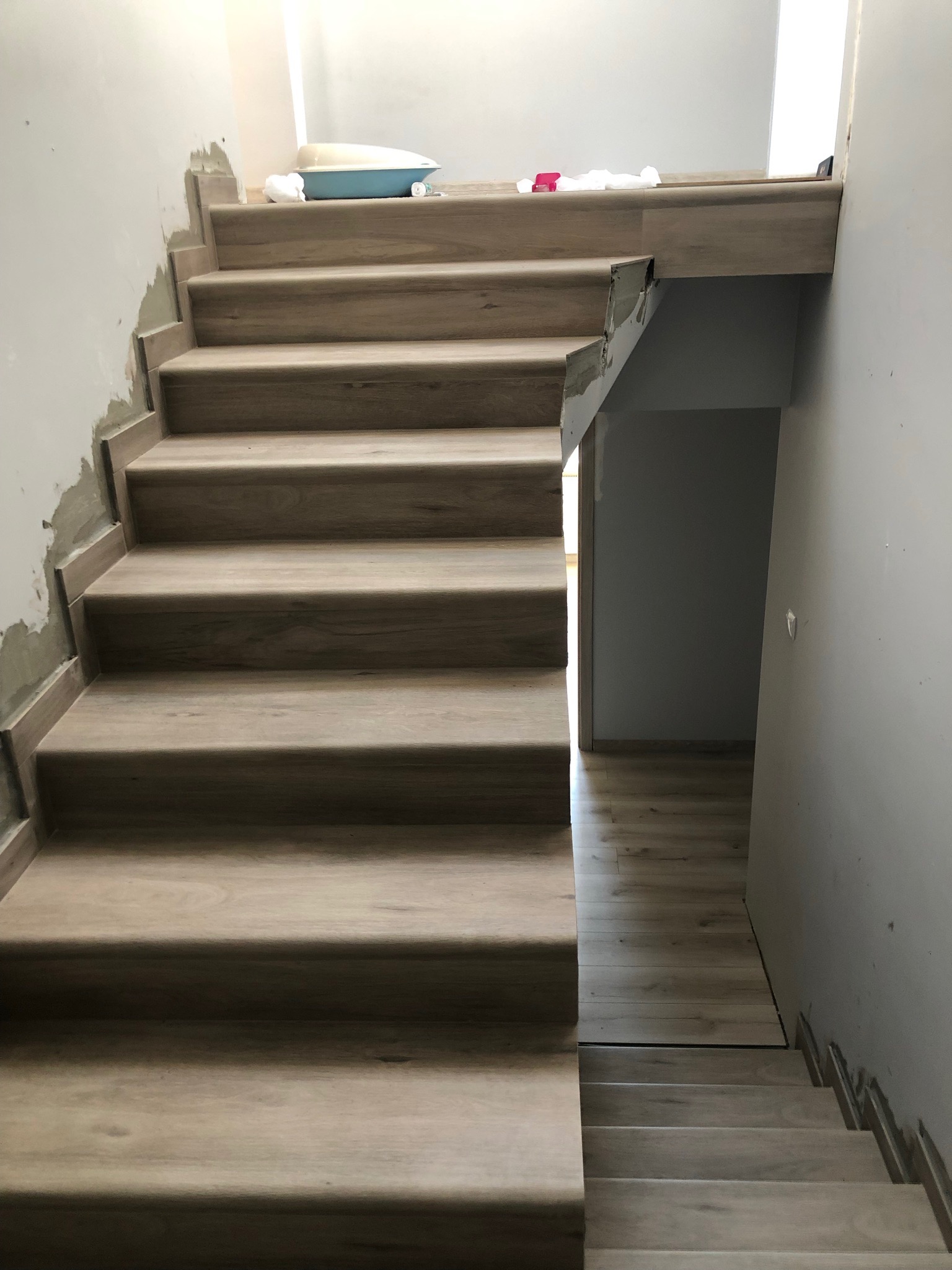 Montaż drewnopodobnych paneli na schodach wewnętrznych, widoczne niedoskonałości w wykończeniu ścian i resztki materiałów budowlanych, pomieszczenie w trakcie prac wykończeniowych.