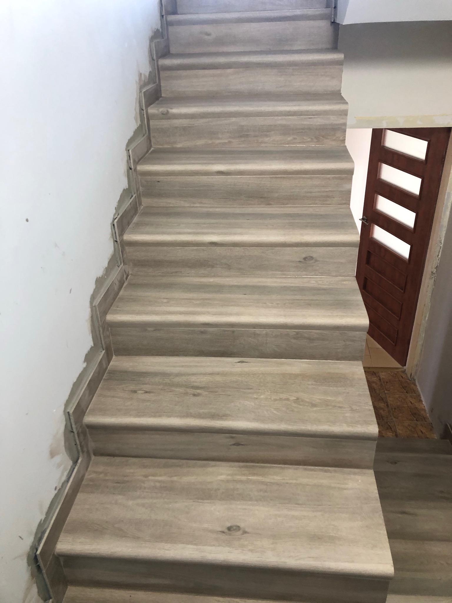 Schody wyłożone panelami imitującymi drewno w trakcie montażu, widoczne niedokończone wykończenia przy ścianie i brązowe drzwi z przeszkleniami w tle.