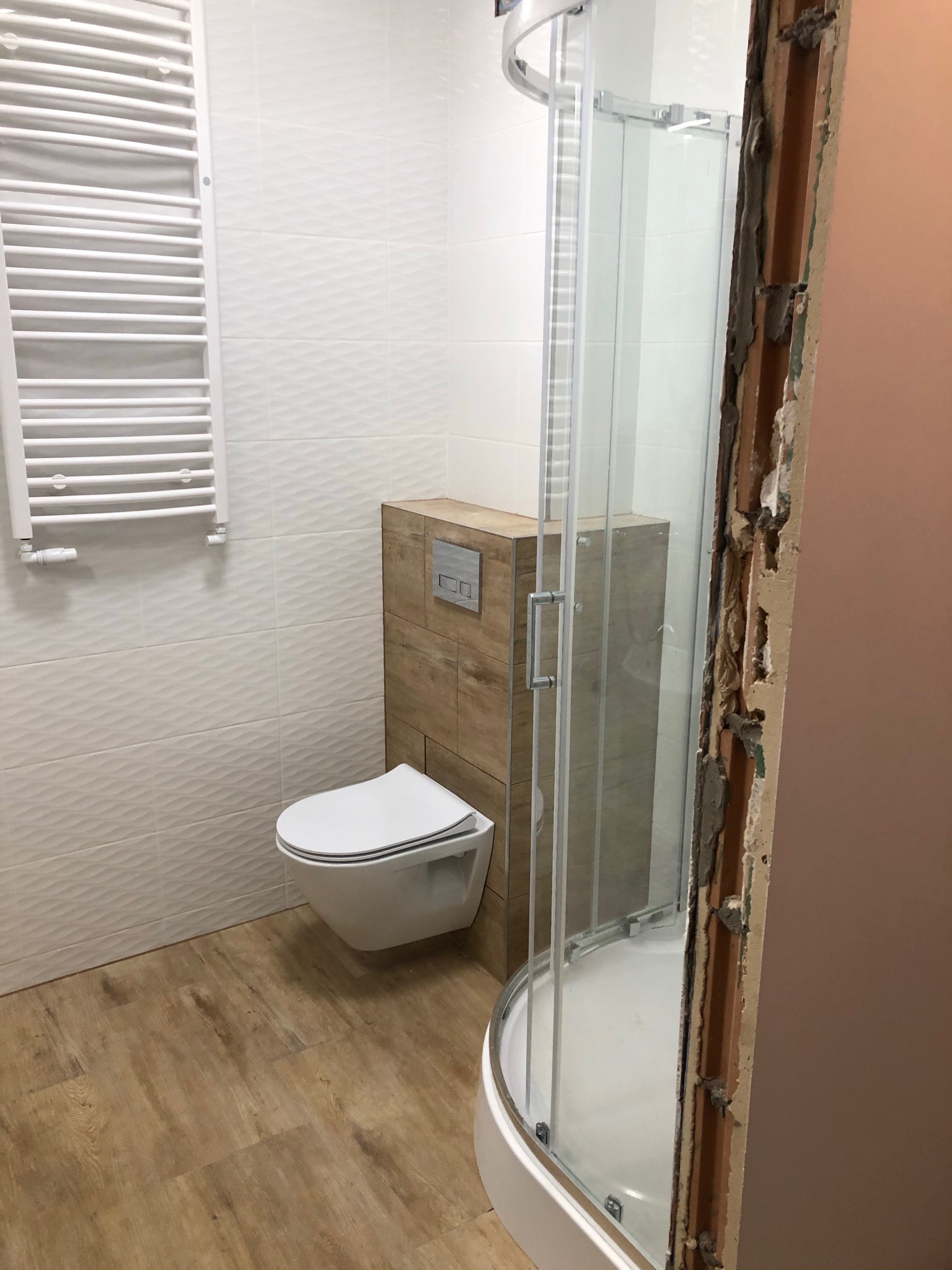 Nowoczesna łazienka w trakcie wykańczania: podwieszana toaleta, grzejnik drabinkowy na ścianie z trójwymiarowymi płytkami, kabina prysznicowa z widocznym surowym obramowaniem.