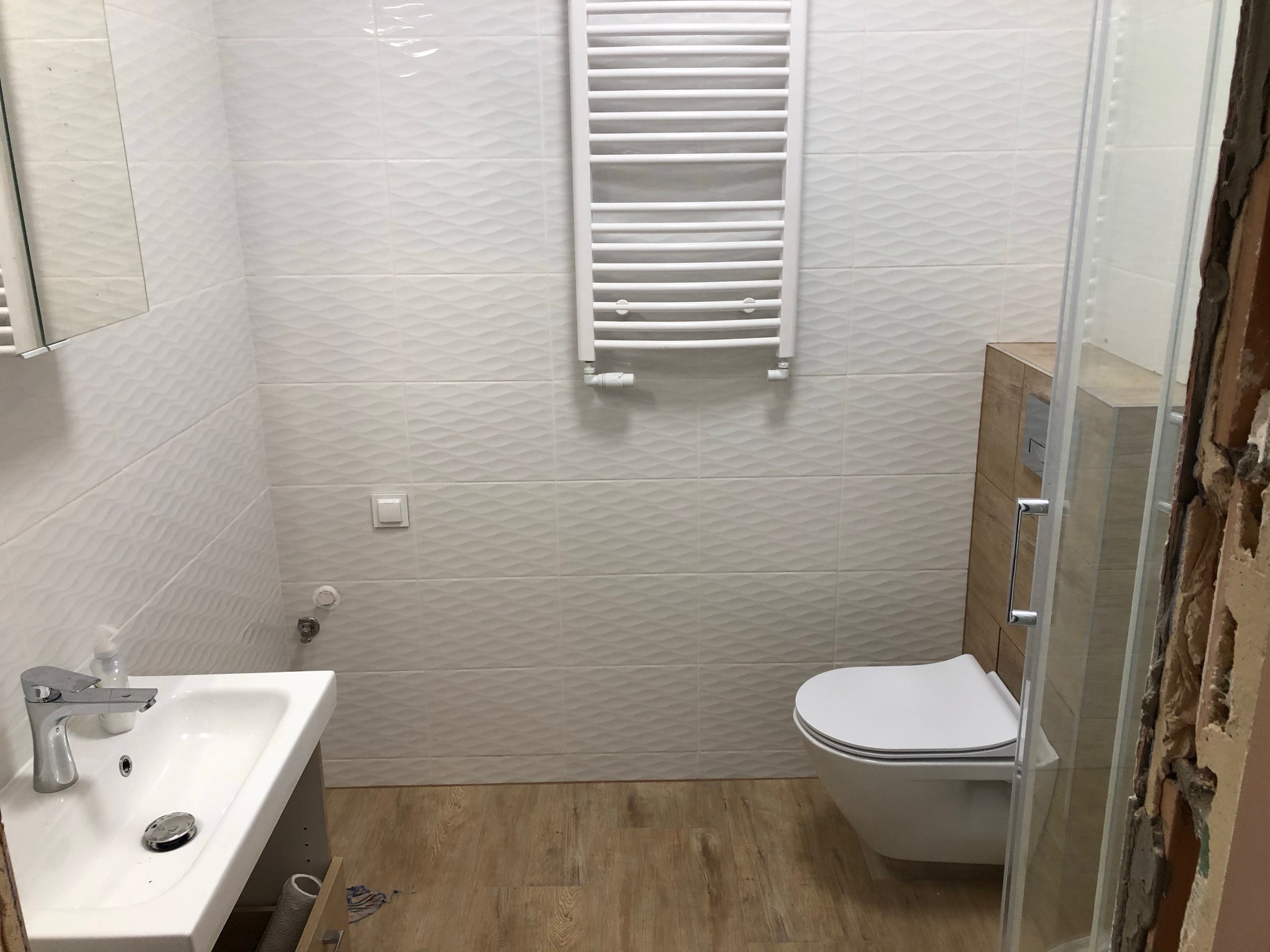 Nowoczesna łazienka po remoncie z umywalką, toaletą wiszącą i grzejnikiem drabinkowym na ścianie z białych płytek o falistej strukturze, widoczny fragment kabiny prysznicowej w trakcie montażu.