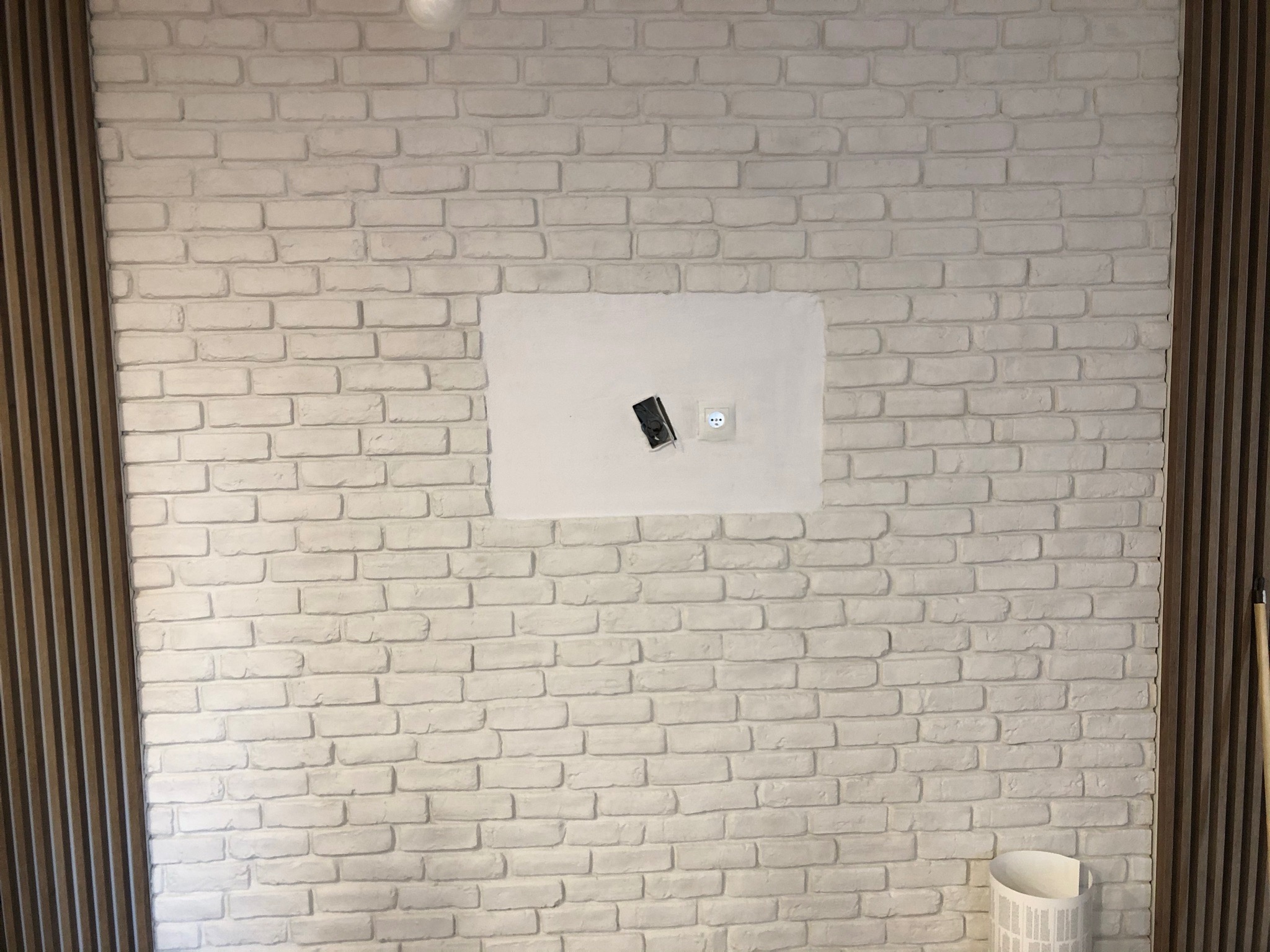 Ściana z białej cegły dekoracyjnej z drewnianymi panelami po bokach, wnęka z gładzią, gniazdkiem i puszką elektryczną, widoczny fragment białego kosza na śmieci u dołu.