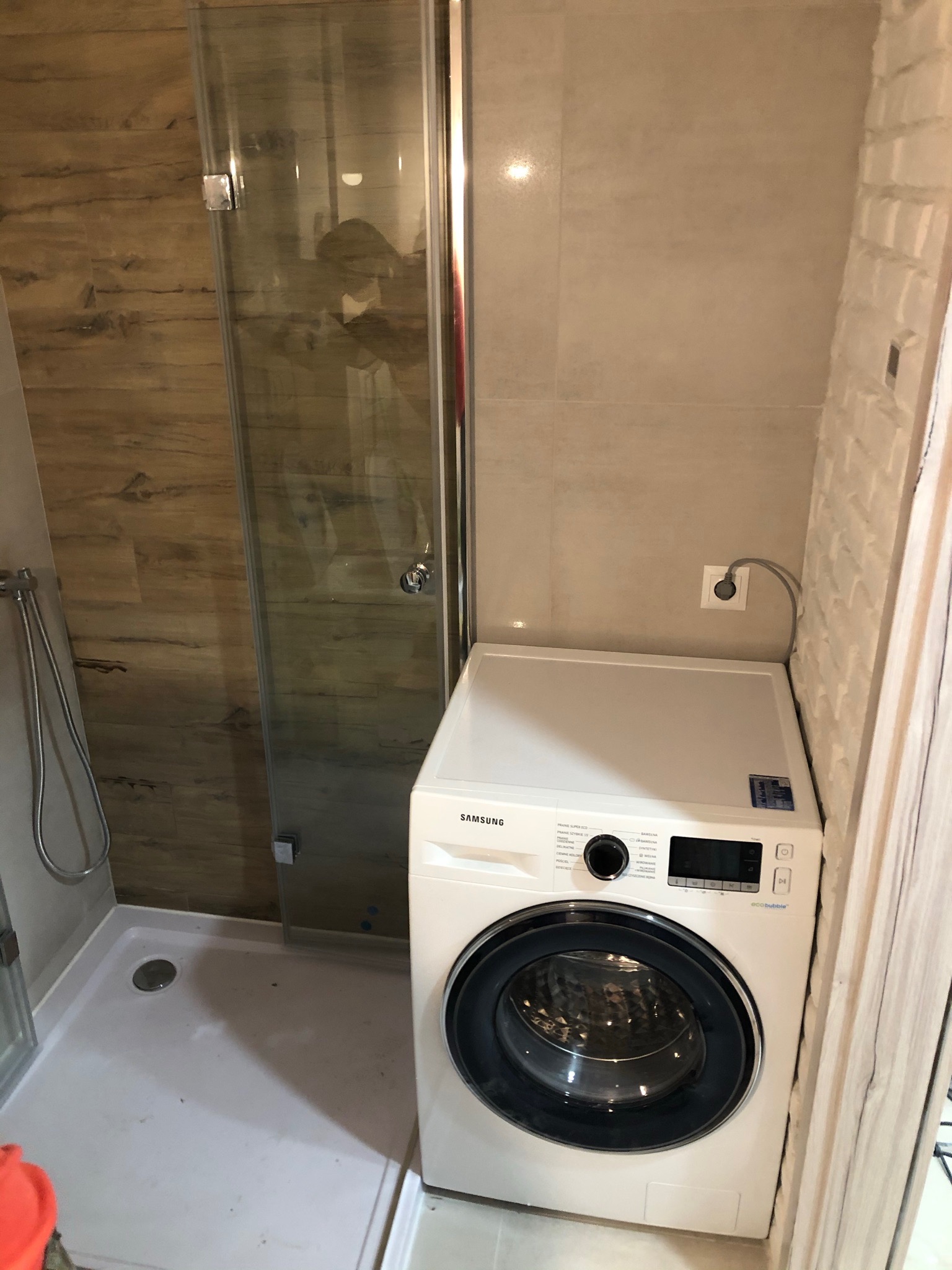 Wąska łazienka z kabiną prysznicową z chromowanymi okuciami i pralką Samsung, ściany wyłożone płytkami imitującymi drewno i cegłę.