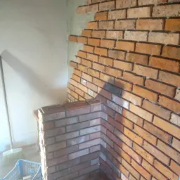 Fragment ściany z cegły klinkierowej w trakcie montażu, widoczne niedokończone fragmenty, nieotynkowana powierzchnia i niebieski pojemnik z narzędziami w tle.