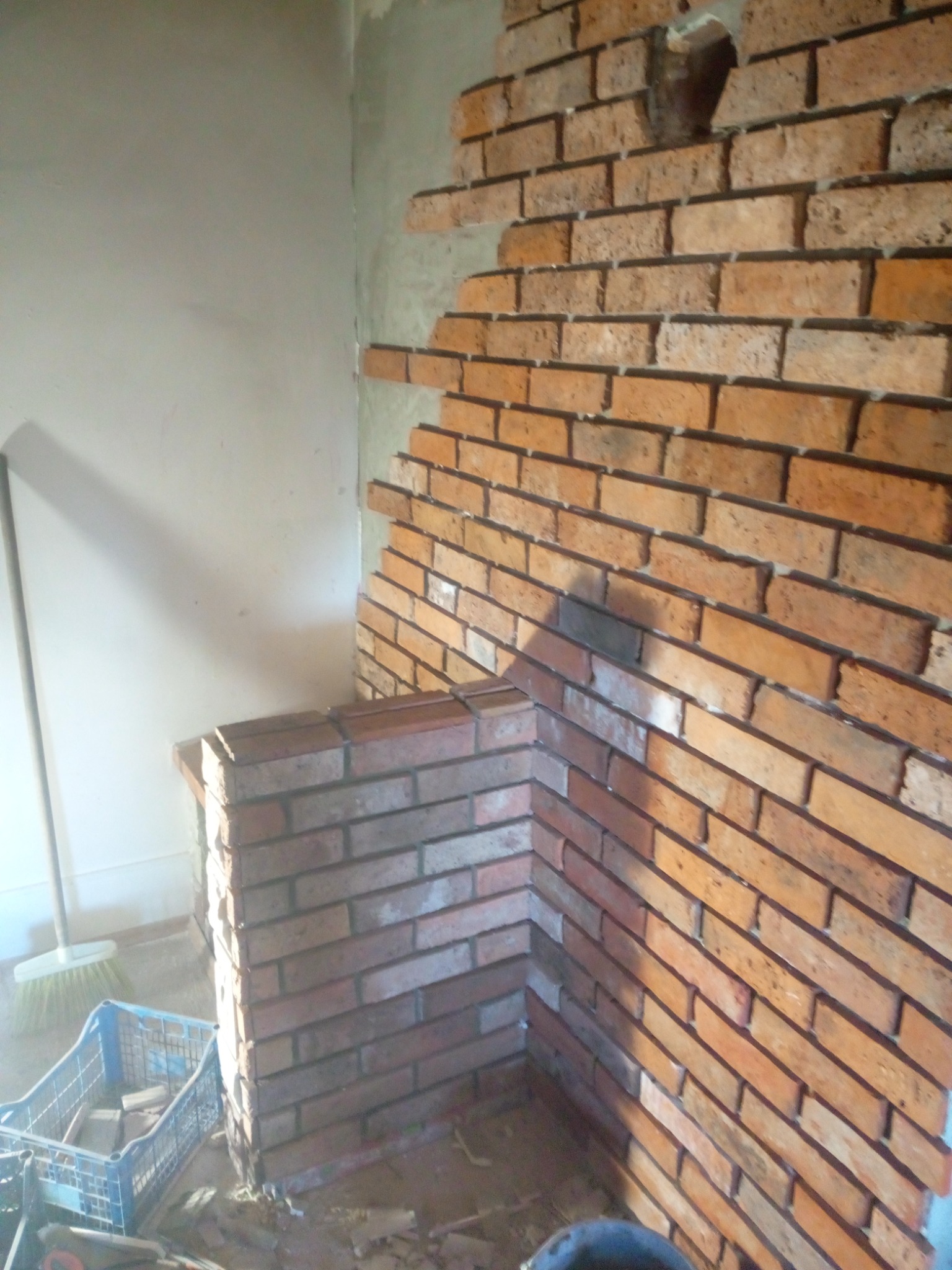 Fragment ściany z cegły klinkierowej w trakcie montażu, widoczne niedokończone fragmenty, nieotynkowana powierzchnia i niebieski pojemnik z narzędziami w tle.