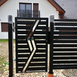 Czarna, nowoczesna brama wjazdowa z poziomymi panelami i geometrycznym wzorem w kształcie rombu, częściowo owinięta folią ochronną na słupkach, osadzona na żwirowym podłożu przed białym domem...