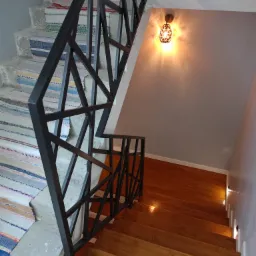 Wnętrze domu z drewnianymi schodami i czarną, geometryczną konstrukcją zabezpieczającą, częściowo wykończone schody betonowe z kolorowymi chodnikami, oświetlenie punktowe wzdłuż ściany.