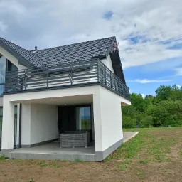 Dwukondygnacyjny dom z płaską, metalową balustradą na balkonie i tarasem z szarymi meblami ogrodowymi, widok z zewnątrz na tle zachmurzonego nieba i zieleni.