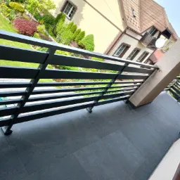 Czarna, pozioma balustrada z metalowych profili na balkonie w Łabowej, z widokiem na ogród i budynek z beżową elewacją.