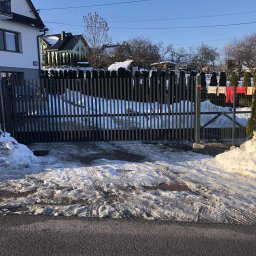 Stalmar - Nowoczesna, grafitowa brama wjazdowa z pionowych profili, z widocznymi śladami opon na śniegu i topniejącym śniegiem po bokach, na tle domów i zielonych tuj.