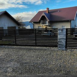 Stalmar - Nowoczesna brama wjazdowa z poziomych profili, osadzona na kamiennych słupkach z wbudowaną skrzynką na listy. Tło stanowi dom i malowniczy krajobraz.