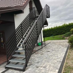 Stalmar - Zewnętrzne schody metalowe z brązową balustradą, prowadzące do drzwi na piętrze domu. Schody z ażurowymi stopniami, ułożone na kostce brukowej. Widok z dołu, pochmurne niebo.