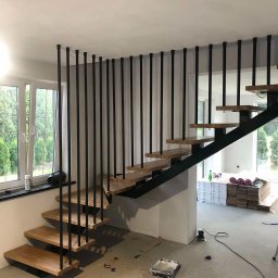 Stalmar - Nowoczesne schody metalowe z drewnianymi stopniami i czarną balustradą z pionowych prętów. Widok na okno i wnętrze domu. Podłoga w trakcie wykończenia.